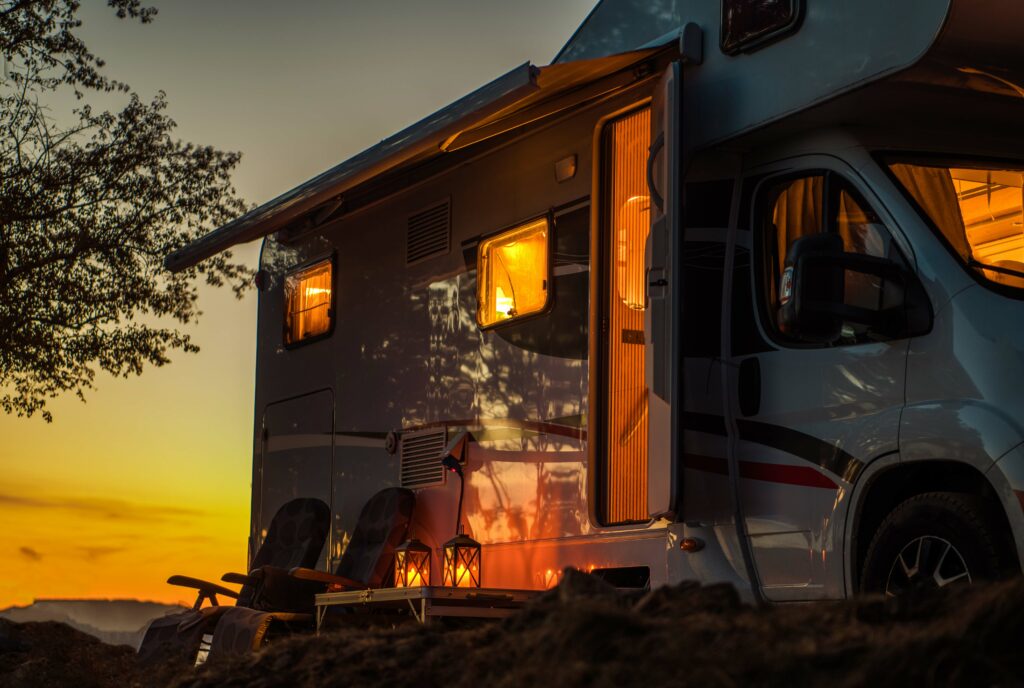An RV at dusk with warm light spilling out. There are two camp chairs set up out in front of it.