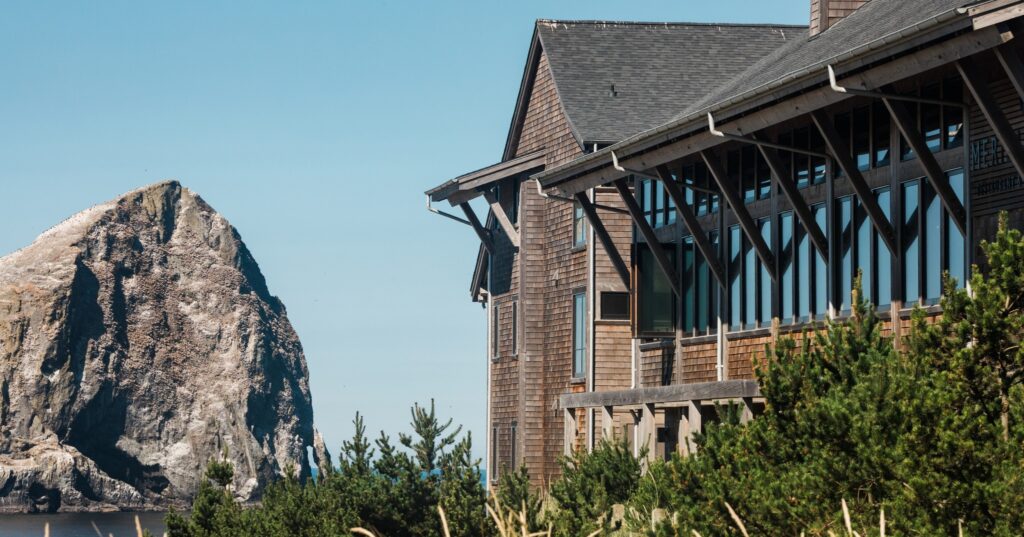 The outside of the lodge in front of Haystack rock. The lodge has a black roof and wood siding.