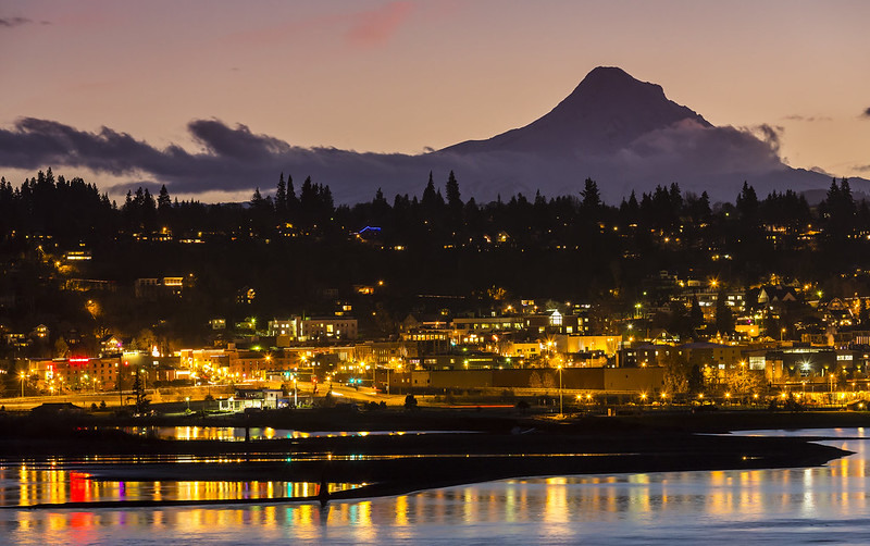 A night time view of Hood River, Oregon as seen from across the Columbia River. Lights reflect off of the river and Mount Hood looms in the background silhouetted against the late evening sky, best oregon towns, spring road trip, best towns to visit, 2024