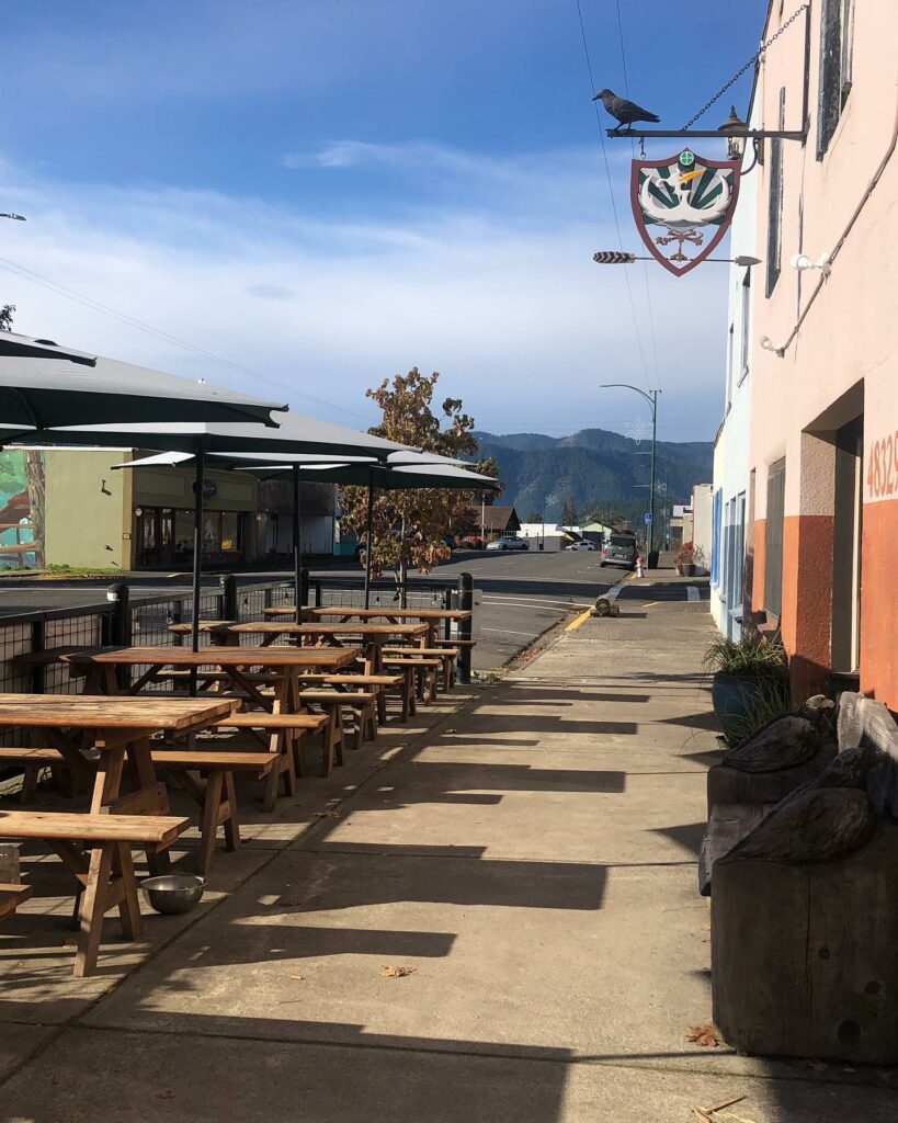The outside of The 3 Legged Crane in Oakridge, Oregon. There are picnic tables under umbrellas on the sidewalk, and a view of the forested mountains in the distance, best oregon towns, spring road trip, best towns to visit, 2024
