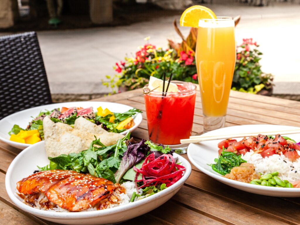 Three plates of colorful seafood and a tall orange drink with a shorter red drink on an outdoor wooden table at Anthony's.