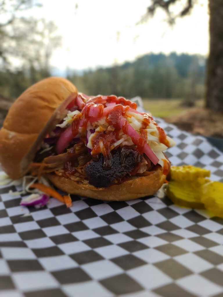 A pork sandwich with the top bun sitting off to the side. It has colorful onions on top and pickles sitting on the side. It's sitting on a black and white checkered paper food wrapper.