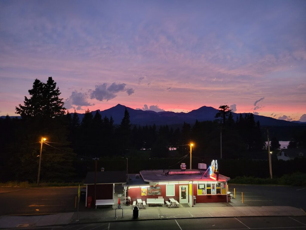 Eastwind Drive-In all lit up at night as the last of the light disappears over nearby mountains silhouetted in the distance, fringed in pink light.